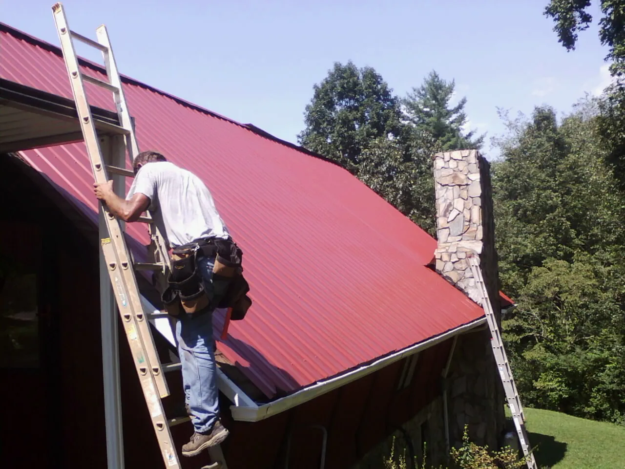 Roofer on a ladder beside a red metal roof and stone chimney — Soffit & Fascia Repair by Roofing Brothers in Springer